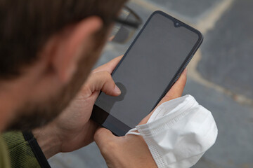 A young man, grabbing a protective face mask, holds a smart mobile phone with his hand, Spain.