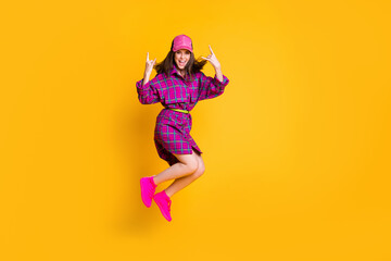 Full size photo of brunette hair lady jump show rock sign wear checkered cap outfit isolated on yellow color background