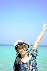 Asian women(half body) smiling happily raising their arms, wearing a floral blouse, woman hat with black ribbon,background is blue sky and a light green sea, holiday travel concept,with copy space.
