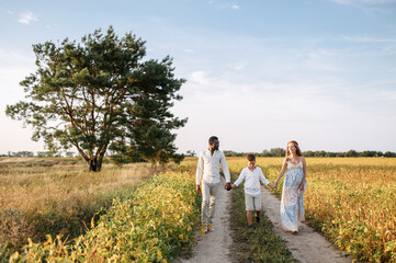 Multiracial family on the meadow in the summer day. A pregnant wife, a black husband and school age son walking holding hands
