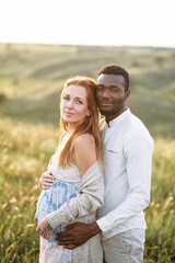 Relaxed multiracial pregnant couple in the park outdoor. An african american husband hugs from the back his charming pregnant wife