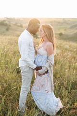 Fototapeta premium Stylish diverse couple. An african-american husband and his caucasian pregnant wife are holding hands and hugging outdoors