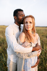 Summer love story on the meadow. An african american man in hugging charming pregnant young woman with care and awe