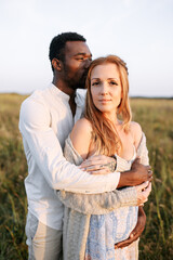 Obraz premium Summer love story on the meadow. An african american man in hugging charming pregnant young woman with care and awe