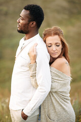 Obraz premium Multiracial couple in love in sunset light. A charming red haired woman with eyes closed hugging from the back an african-american man