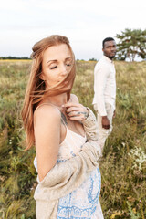 Beautiful red haired pregnant woman in rustic style sundress on the meado, her husband in out of focus on the background
