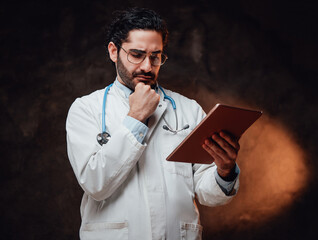 Holding a tablet in dark background skilled doctor dressed in white coat with glasses and serious face.