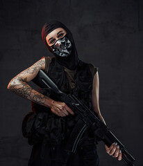 Tattooed and joyful woman in black armour and weared with mask poses in dark background holding assault rifle.