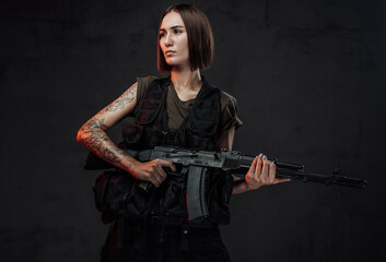 Dressed in black armour with dark shirt martial woman with short haircut poses in dark background holding assault rifle.