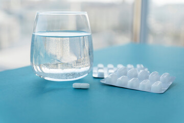 transpatent glass with pure water and white pills in blister on blue table near window, copy space for text. Treatment from headaches, flu, influenza, viruses. Healthcare concept