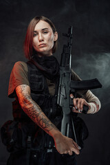 Fototapeta premium Seductive female soldier with tattoo and bandaged hand poses in dark foggy background holding rifle on her shoulder.