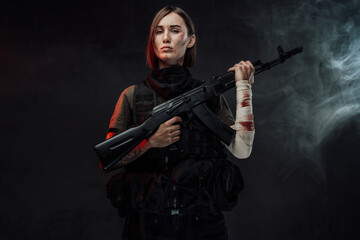 Fototapeta na wymiar Dressed in dark armour military woman with short haircut and bandaged hand holds her assault rifle in dark smokey background.