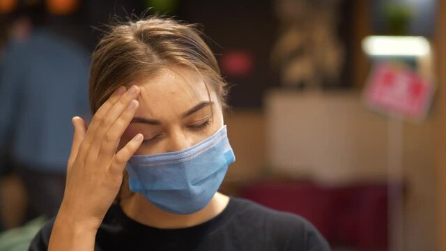 Sick young girl in blue medical protective mask inside a restaurant, she is not feeling well and has a headache, she massaging head