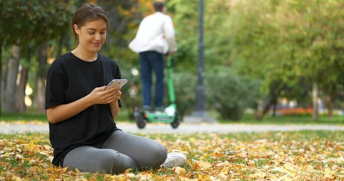 Young beautiful girl sits on lawn in autumn city park, uses smartphone, looks and flips through the photos, smiles and laughs. Two guys on electric scooters drive along road. Cinema 4K 60fps video