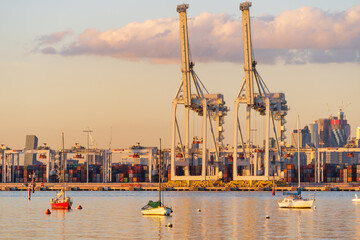 Small boats anchored in front of large gantry cranes at a city port at sunset.
