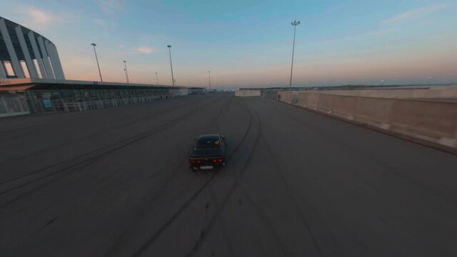 Black sports jdm car drives along empty ground and does drift tricks against city under dark blue sky in evening aerial first point view. Fpv freestyle cinematic shot.