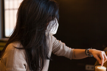 Fototapeta premium Adult spanish woman sitting on a coffeeshop wearing protective mask during pandemics stirring his coffee while speaks with someone with natural light and black negative space.