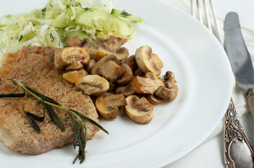 On a white plate lies aromatic grilled meat with rosemary and mushrooms and salad with vegetables.