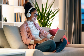 Beautiful young entrepreneur woman working with laptop while listening music with headphones sitting on couch in the office. - Powered by Adobe