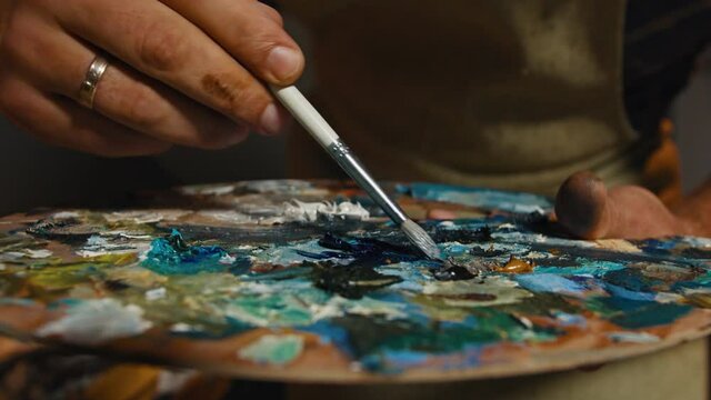 Painter is taking colors by paintbrush from palette. Close-up paints in art workshop during masters work. Art therapy, fine art concept