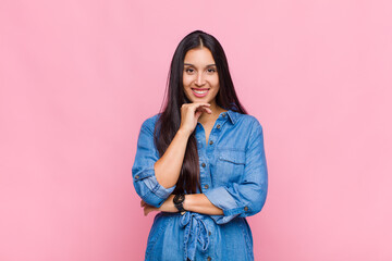 young woman looking happy and smiling with hand on chin, wondering or asking a question, comparing...