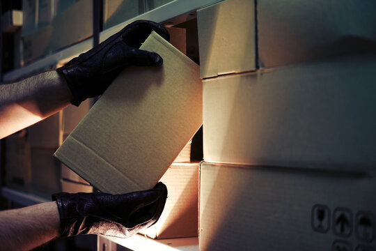 Thief hands with gloves steal a box of goods in a warehouse in the dark. Concept of problems with theft of goods and postal parcels