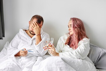 sick male has temperature and headache lying on bed, woman supports him, give medications