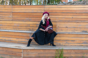 Cheerful sits on a summer theater bench, wooden in a burgundy coat and biret, with a charismatic appearance, in the fall against the background of trees a blue sky.