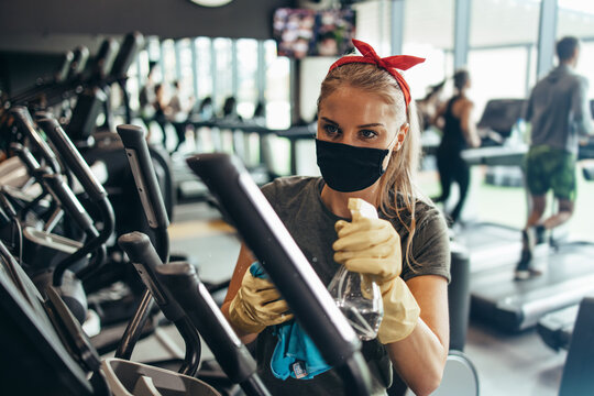 Young female worker disinfecting cleaning and weeping expensive fitness gym equipment with alcohol sprayer and cloth. Coronavirus global world pandemic and health protection safety measures. - Powered by Adobe
