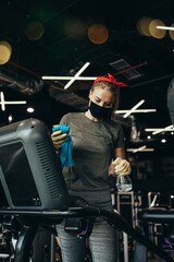 Obraz na płótnie Canvas Young female worker disinfecting cleaning and weeping expensive fitness gym equipment with alcohol sprayer and cloth. Coronavirus global world pandemic and health protection safety measures.