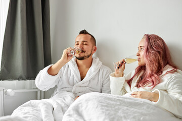Obraz na płótnie Canvas lovely woman and man in white robes lying on bed and enjoying time together, drinking beverage together, love, weekends, rest concept