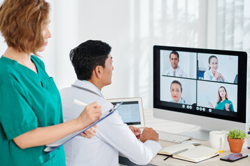 Chief physician having online conference with colleagues when his assistant taking notes in documents