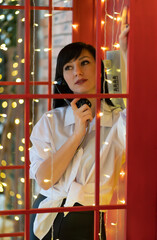 A brunette woman stands in a red phone booth and talks on the phone