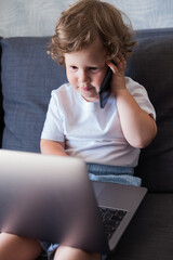 Cute little child is sitting on the sofa with a computer and talking on the phone. Remote work. Working at home on the couch.