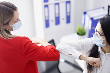 Fototapeta premium Two businesswomen in protective face masks touching elbows in office. Prevention measures during covid-19 pandemic concept
