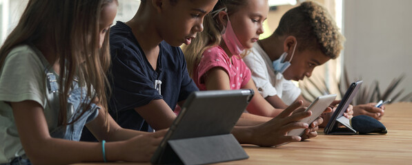 Four diverse kids wear facemasks sit at table use wireless gadgets