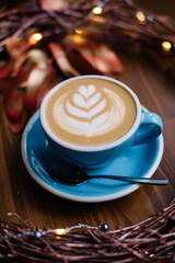 Ceramic blue cup of delicious fresh cappuccino coffee on the rustic wooden background decorated with festive hand made wreath, fireflies and copper painter leaves