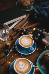 Two delicious cups ofbcappuccino coffee standing on the rustic wooden table among Christmas decorations, fireflies, wrapped gift, scissors, cinnamon sticks, deer toy, with sandwiches on the background