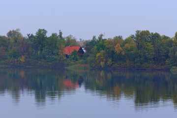 Fototapeta na wymiar Astonishing landscape of autumn colored trees are reflected in the water. Wooden house with red tile roof is hidden in the forest near the river bank. Foggy autumn morning. Kyiv, Ukraine