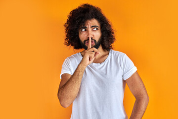 Fototapeta na wymiar young arab man with curly black hair raising his finger on the front of his mouth asking to be quiet, isolated on orange background, he is looking away