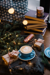 Delicious fresh festive morning cappuccino coffee in a ceramic blue cup on the warm cover with little wrapped gifts, red ornamentals, fireflies and spruce branches