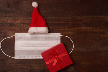 Protective mask and Santa's cap with gift box, flat lay with space for text on brown wooden background