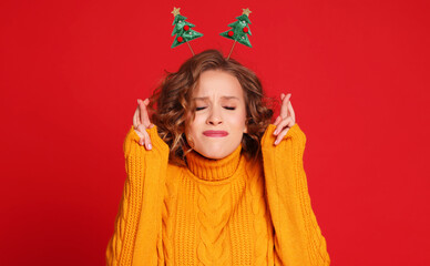 Cheerful woman with christmas headband crosses fingers for good luck, prays and hopes dreams come true, in new year, awaits for miracle  closes eyes on red background.