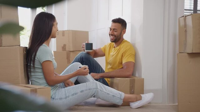 Couple taking a break on moving day sitting on floor surrounded by boxes and making a toast with hot drinks - shot in slow motion