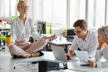 woman sits relaxing while other have brainstorm at work, meditates on table, sits in lotus pose, keeping calm, need some rest