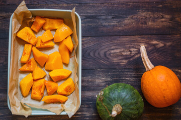 Baked pumpkin sliced in small pieces  in a baking dish on a wooden background with different  fresh pumpkins. Selective focus, copy space. Vegan healthy food.