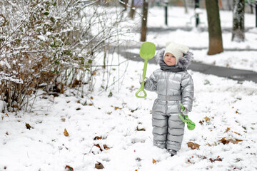 Fototapeta na wymiar Happy child on a winter walk in the park. A funny little girl in a warm silver jumpsuit holds a toy shovel. First snowfall. Dry maple leaves peek out from under the snow. Blurred background.