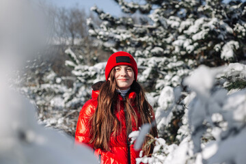 Fototapeta na wymiar Portrait of a girl teenager wearing red coat standing among fir trees covered with snow at the winter park. Young girl having fun in the winter forest. Winter vibes concept.