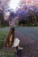A cute little girl with long hair in a white dress stands outside with blooming jacaranda trees