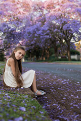 A little cute girl with long hair in a white dress sits on the grass in the street with blooming...